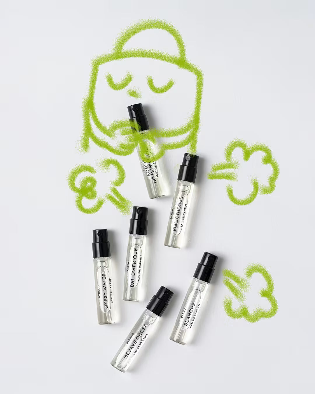 Flat-lay photograph on a white background showing six small Byredo perfume sample spray vials arranged beneath playful green airbrushed cartoon doodles of a character surrounded by cloud-like puffs.