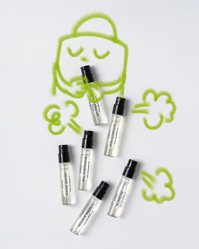 Flat-lay photograph on a white background showing six small Byredo perfume sample spray vials arranged beneath playful green airbrushed cartoon doodles of a character surrounded by cloud-like puffs.